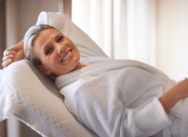 Smiling woman reclining in a white robe, suggesting comfort during a spa or wellness treatment.