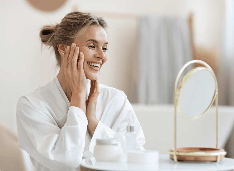 Smiling woman touching her face in front of a mirror, suggesting satisfaction with skincare or self-care routine.
