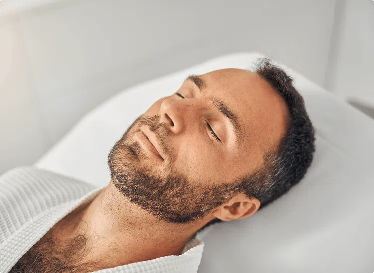 Man lying down with eyes closed in a spa setting, suggesting relaxation or facial treatment.