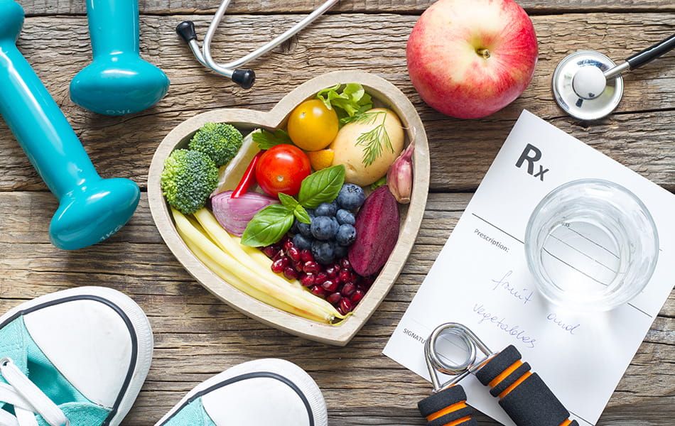 Heart-shaped bowl of fruits and vegetables with dumbbells and prescription, highlighting healthy lifestyle plan.