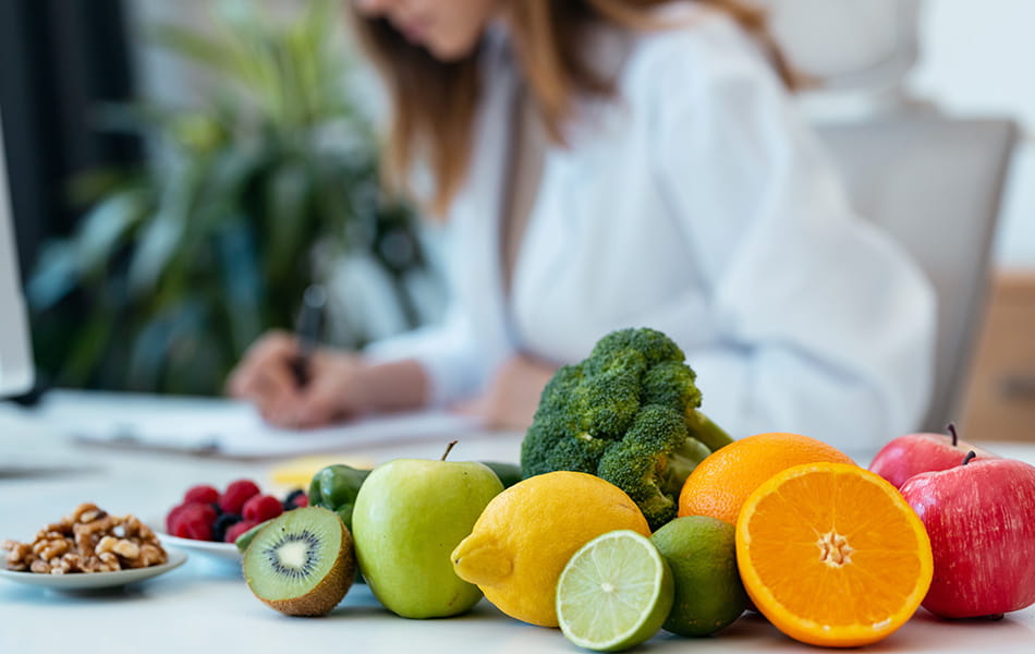 Fresh fruits and vegetables in foreground with woman writing in background, highlighting healthy diet planning.