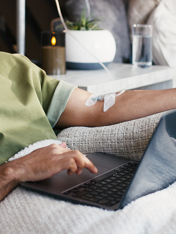 Close-up of arm with IV line while person uses laptop, highlighting convenient at-home infusion therapy.