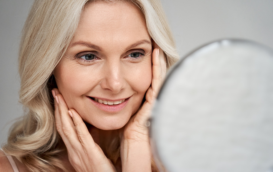 Older woman smiling at mirror with hands on cheeks, highlighting anti-aging skincare results.