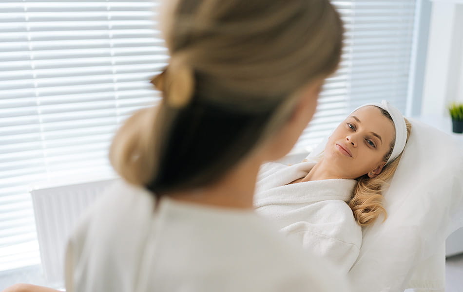 Woman lying in spa bed speaking with esthetician, highlighting facial treatment consultation.
