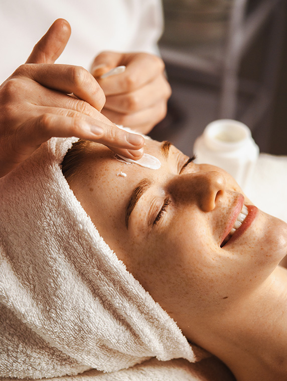 Esthetician applying cream to woman’s forehead during facial, highlighting moisturizing skincare treatment.