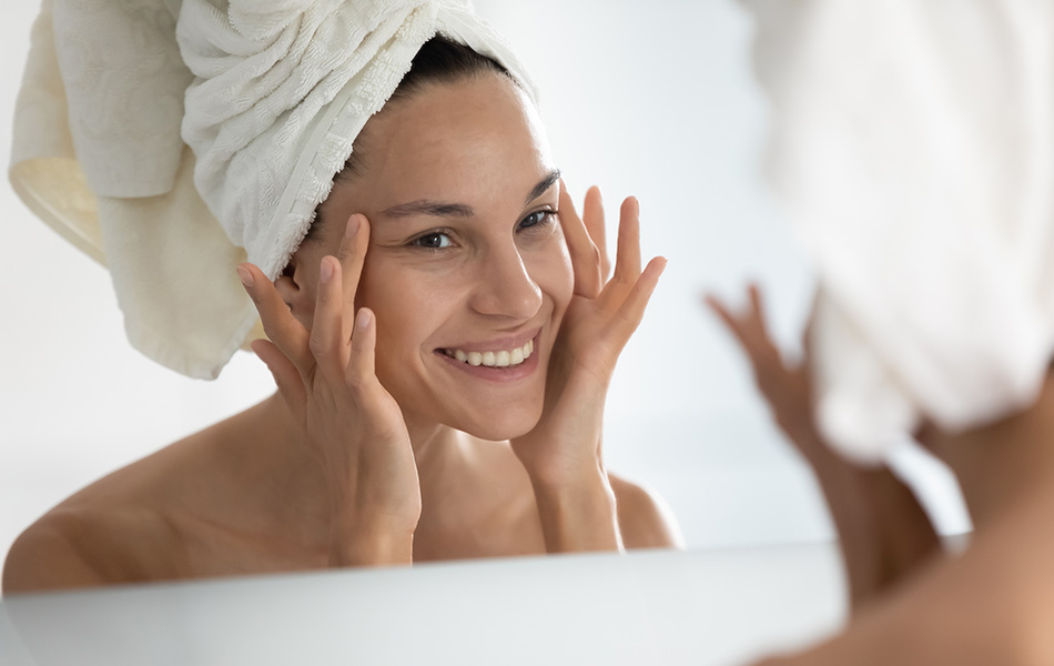 Woman looking at her eyes in the mirror at spa Smiling woman with towel on head touching face in mirror, highlighting fresh skincare routine results.