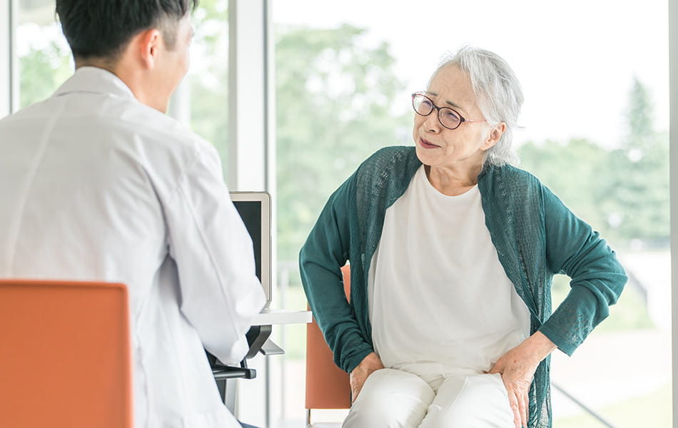 Older woman speaking with doctor and holding lower back, highlighting back pain during medical consultation.